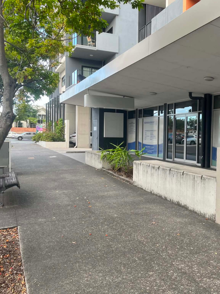 Outdoor walkway to a modern building with glass storefronts and a bench on the left, under green trees