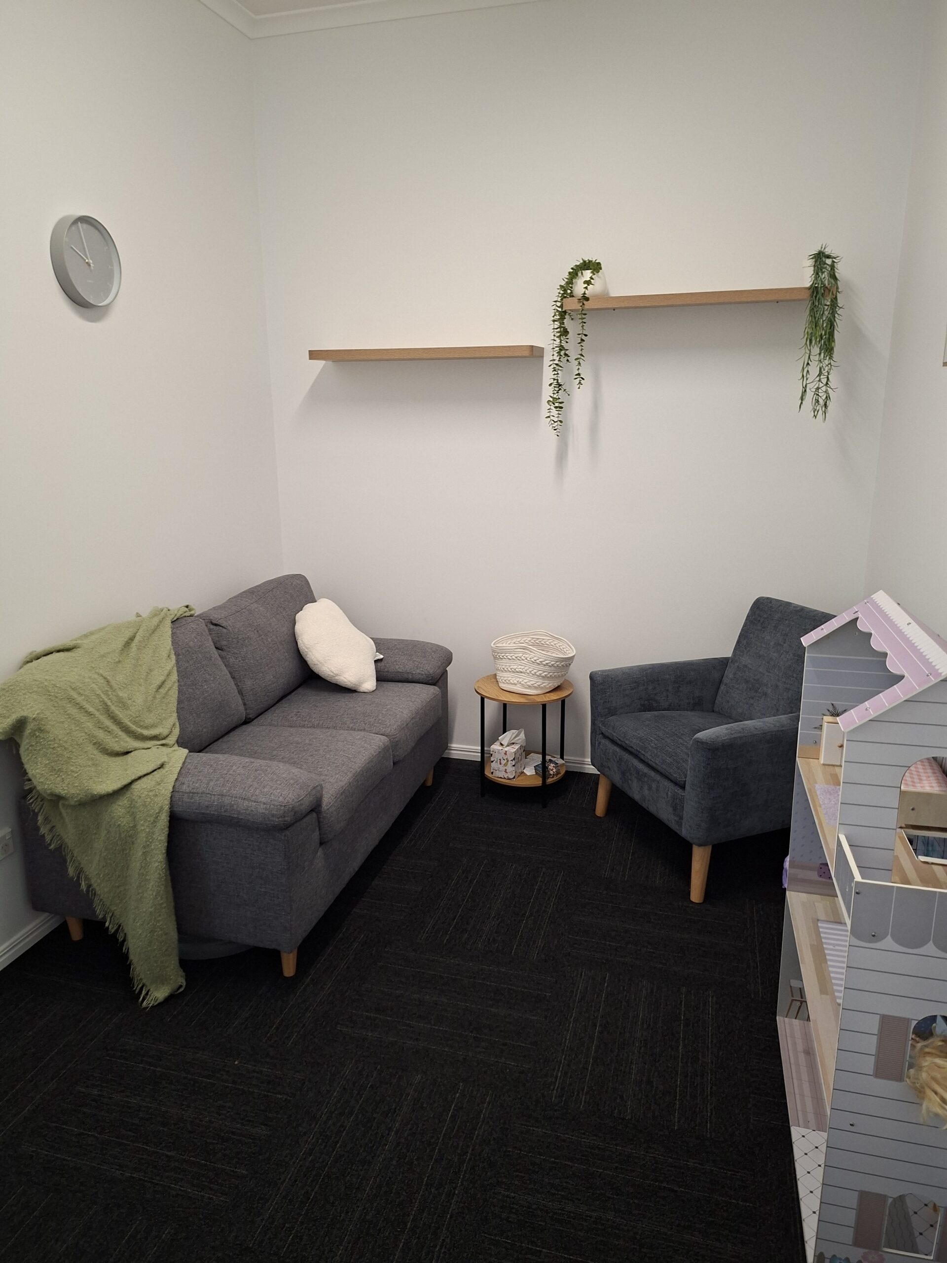 Cozy living room corner with a gray sofa, gray chair, and a small round table topped with a basket next to a plant-lined wall shelf.