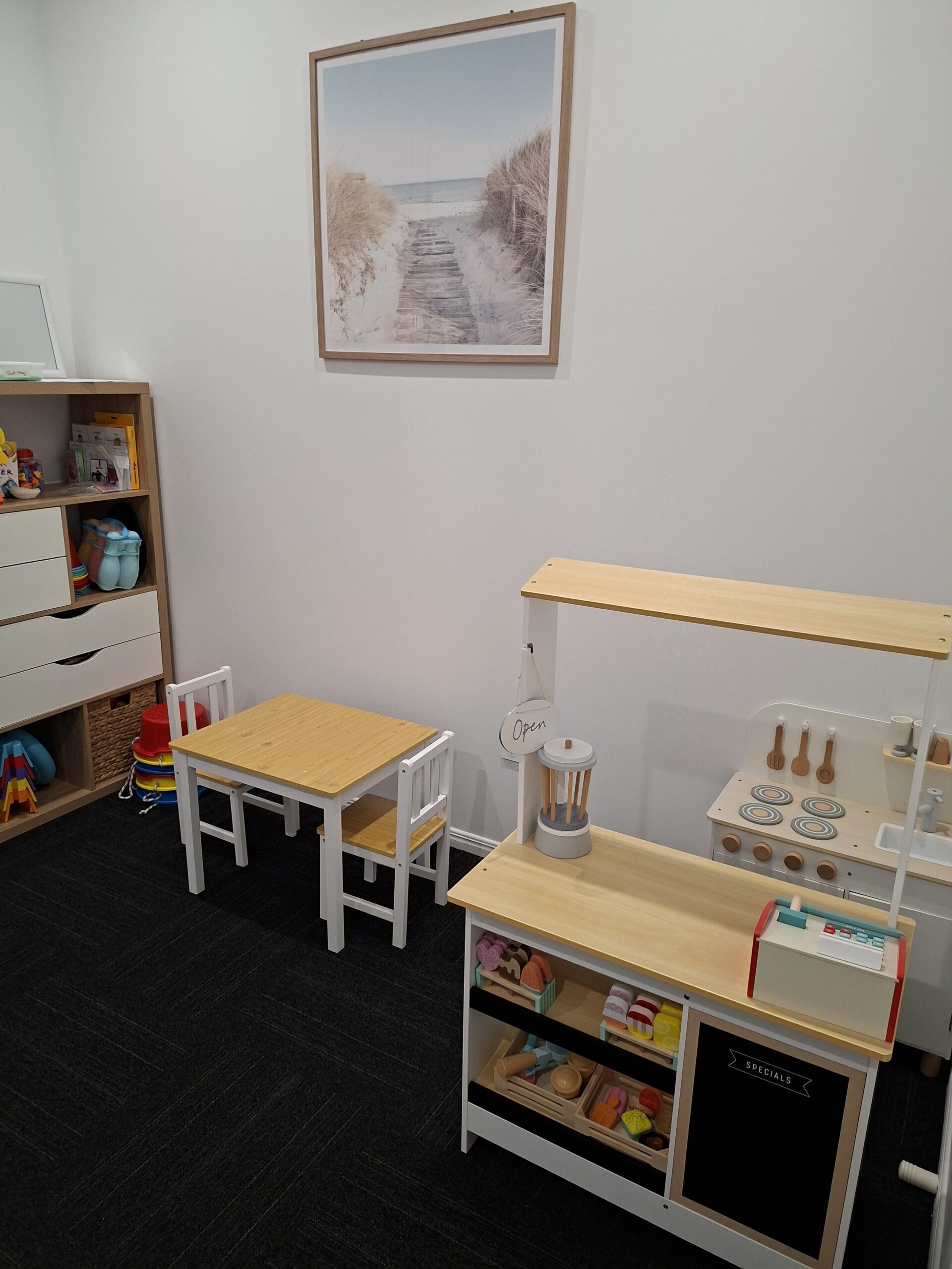 Children's playroom with a white-and-wood toy kitchen on the right, a small wooden table and chairs in the center, a tall shelf of toys on the left, and a framed beach path photograph on the wall.