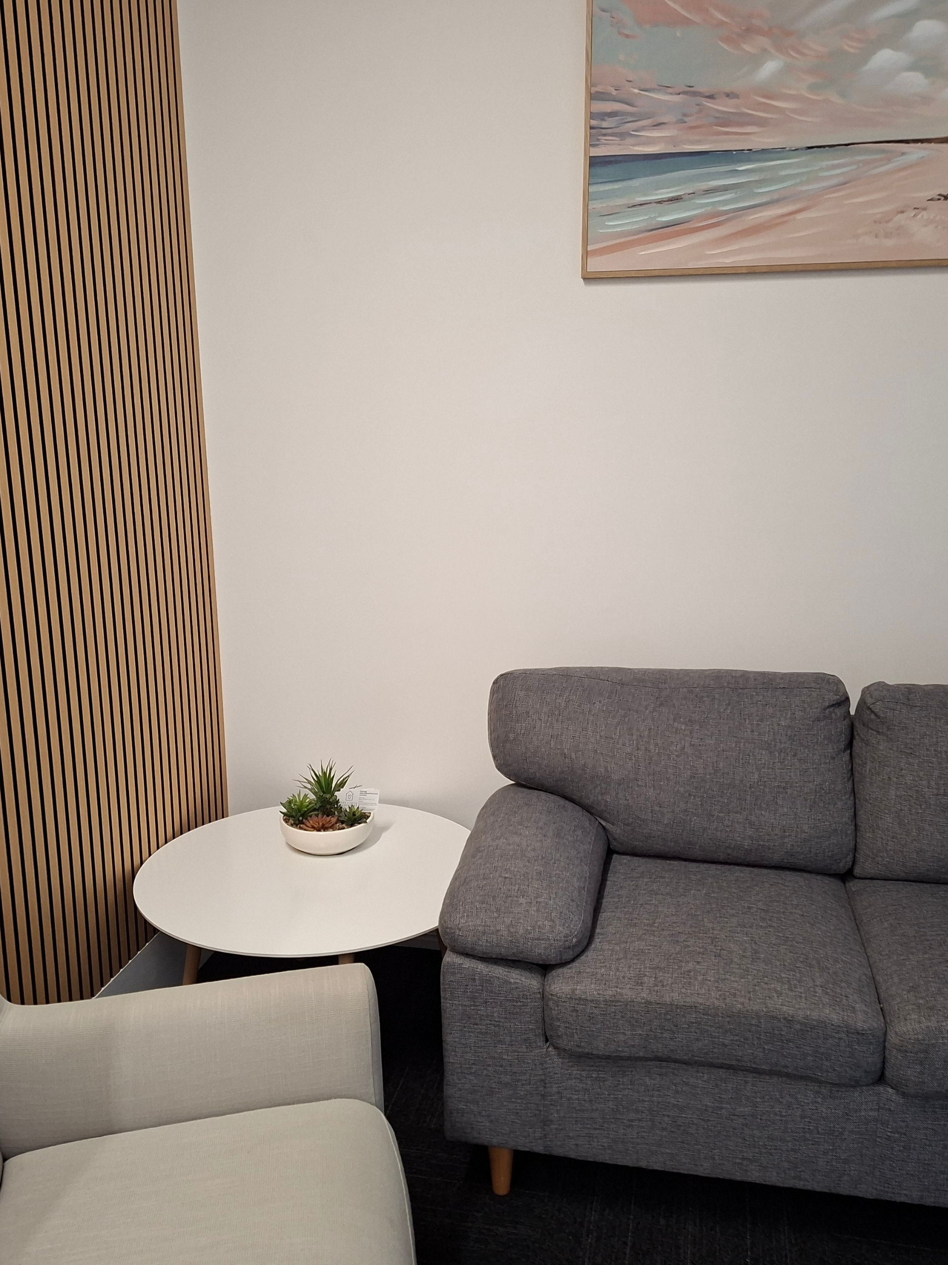 Cozy living room corner with a gray sofa, beige chair, round white table, and a small succulent plant nearby; wooden slat wall on the left and a beach-themed painting above.