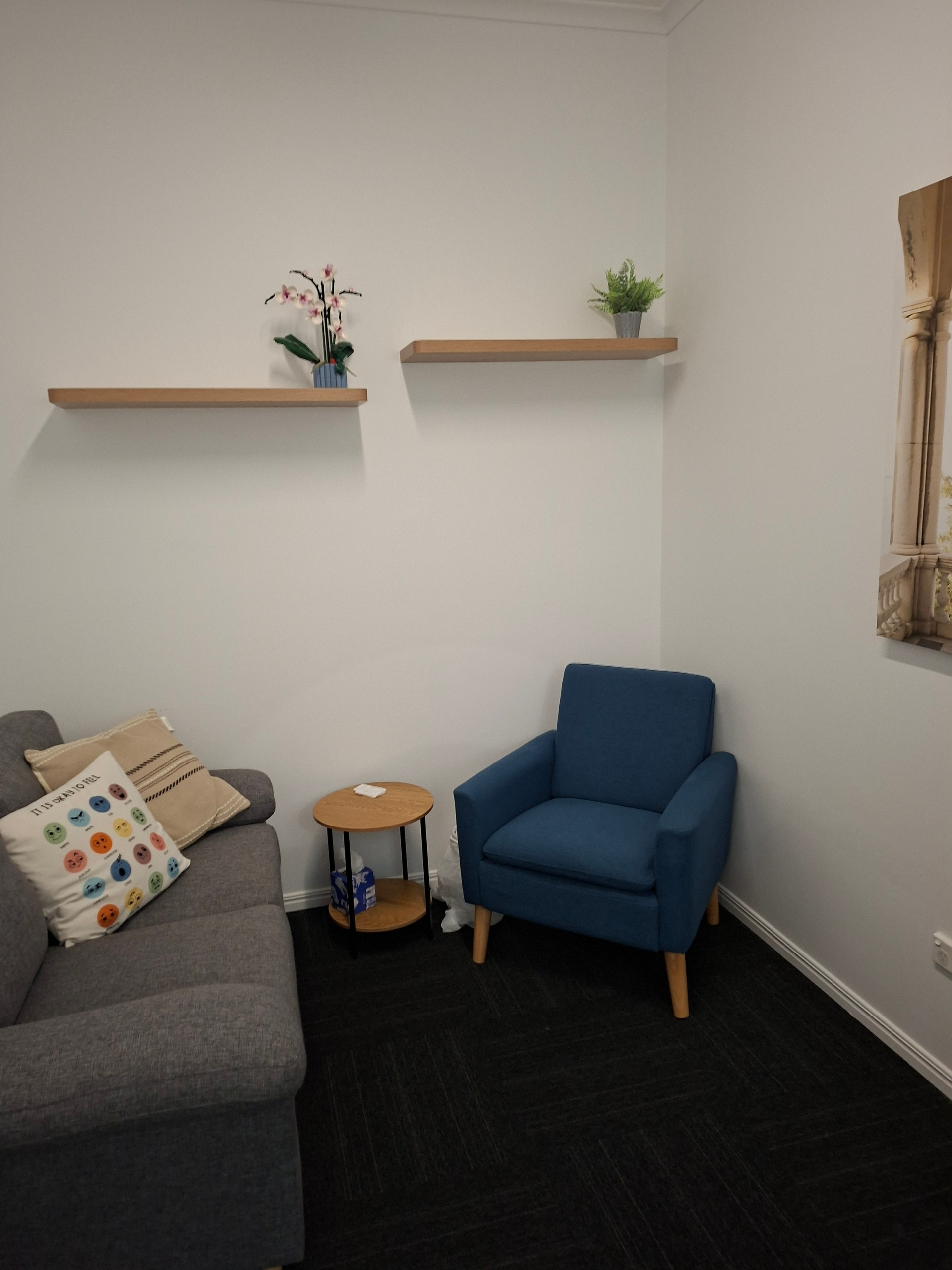 Waiting area with a gray sofa, a blue armchair, and two small round wooden tables in a corner of a room.